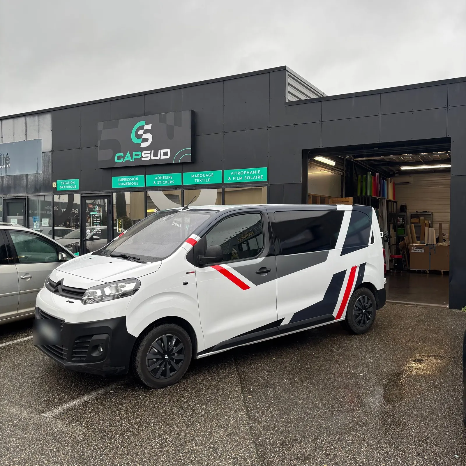 Utilitaire blanc à bandes grises et rouges devant l'atelier